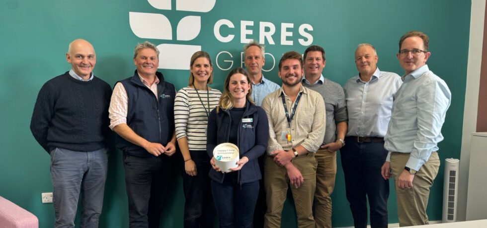 9 people from Ceres Research standing in a row in front of the Ceres Group display wall, one person at the front is holding a small cake that says "happy 1-year anniversary" on the top with the Ceres Research logo.